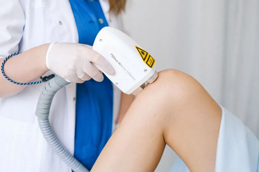 Close-up of a laser hair removal handpiece with a warning label being applied to a client's knee by a practitioner wearing gloves, illustrating the in-clinic, practitioner-led process behind a safety-focused hook