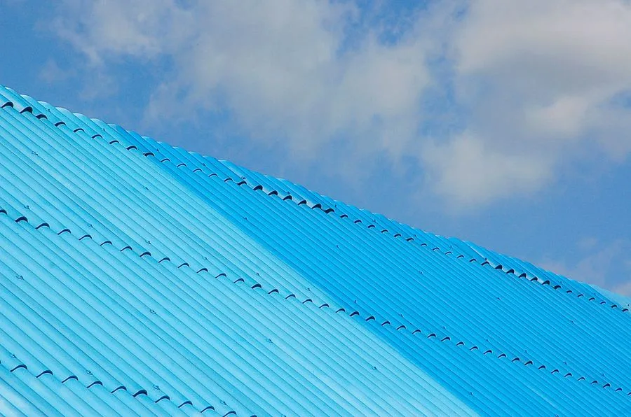 Close-up diagonal view of a blue corrugated metal roof panel against a partly cloudy sky, an example of the metal roofing material and clean installation lines worth showcasing in a before-and-after Instagram post
